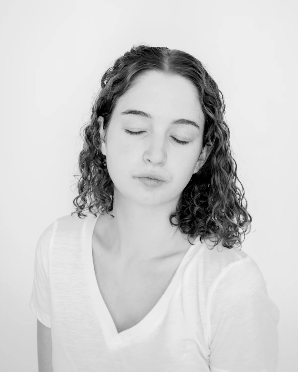 Mulher de olhos fechados, cabelo cacheado e camiseta branca, sobre fundo claro.